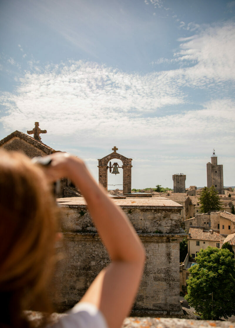 Personne photographiant un clocher avec une cloche à Uzès, vue sur les toits et le ciel légèrement nuageux.